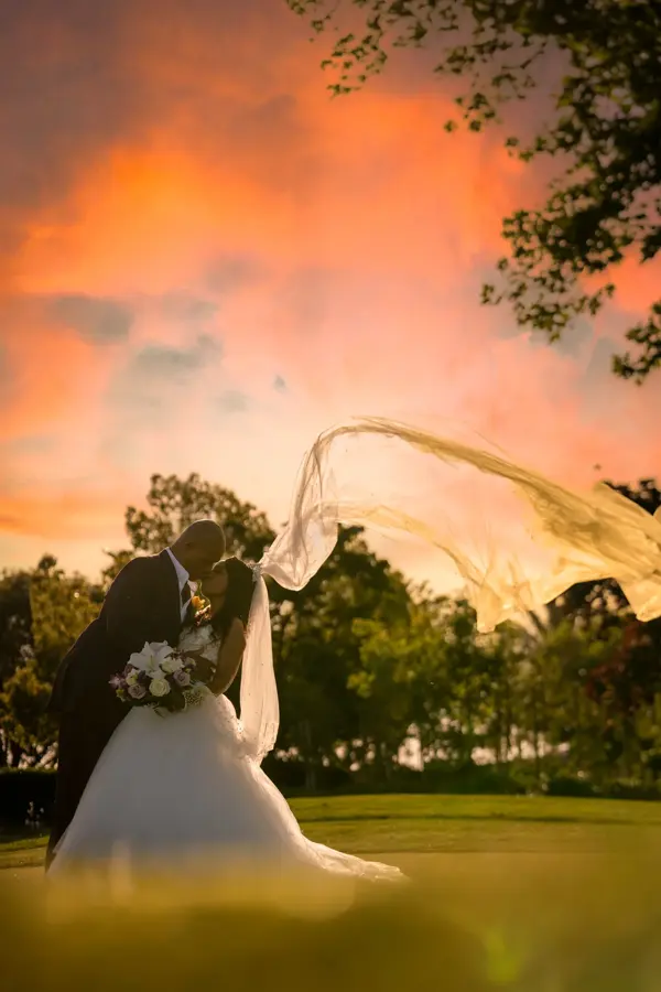 Veil flying against a vivid sunset while the couple kisses